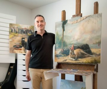 Painter Steve Volpe in his studio next to a work starring a kangaroo in a party hat, called Birthday, and below it, a beach scene, Man and Pyramid. Photo by Rosemary Hasner / Black Dog Creative Arts.