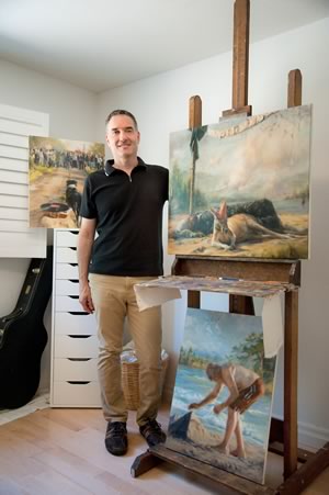Painter Steve Volpe in his studio next to a work starring a kangaroo in a party hat, called Birthday, and below it, a beach scene, Man and Pyramid. Photo by Rosemary Hasner / Black Dog Creative Arts.
